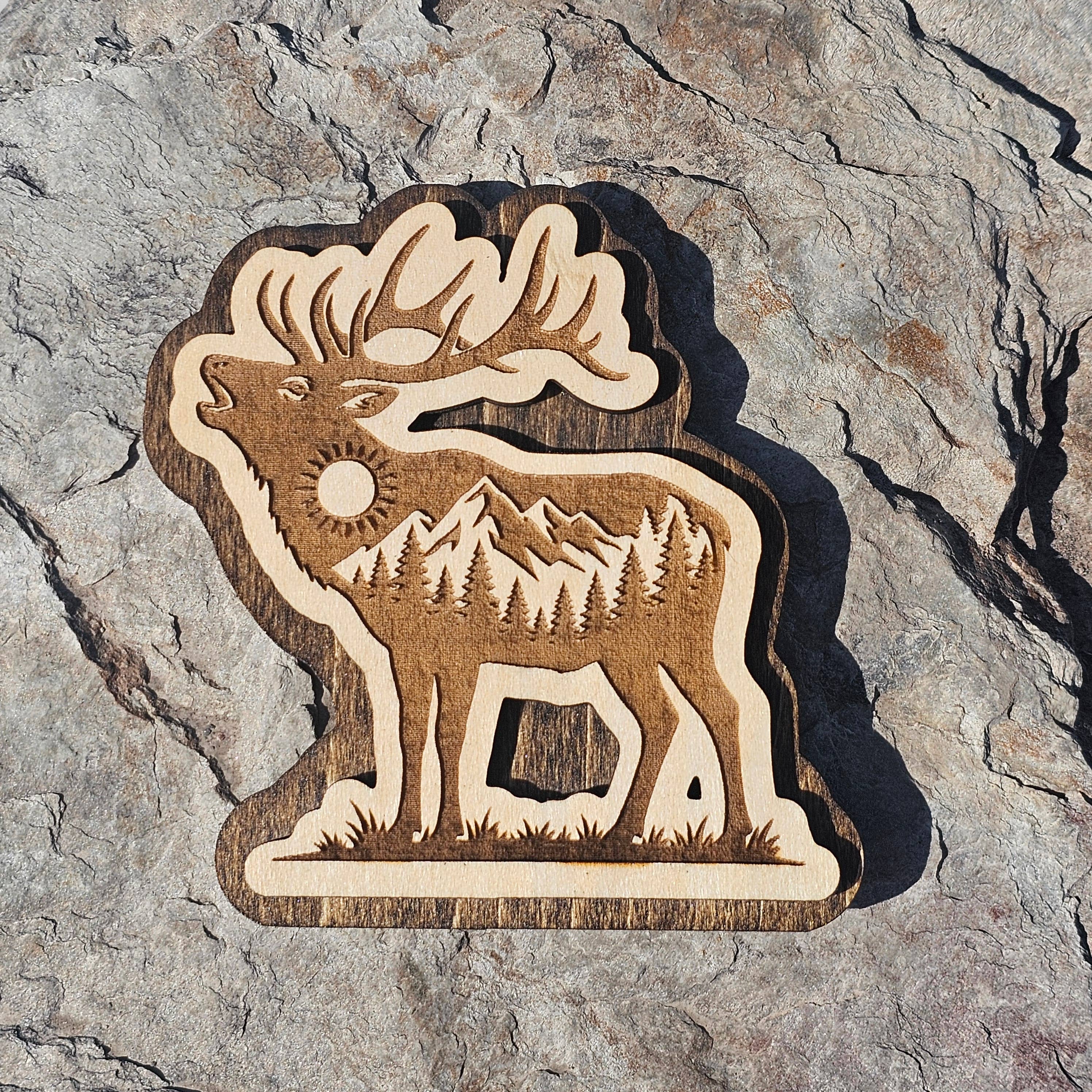 Layered wood magnet of a bugling elk with mountains, pines, and sun engraved inside the silhouette, from The Ruddy Duck in Whitefish, Montana