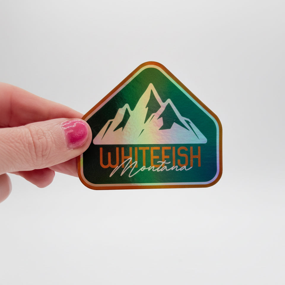 Sticker with mountain design and 'Whitefish Montana' text held by a hand on a white background