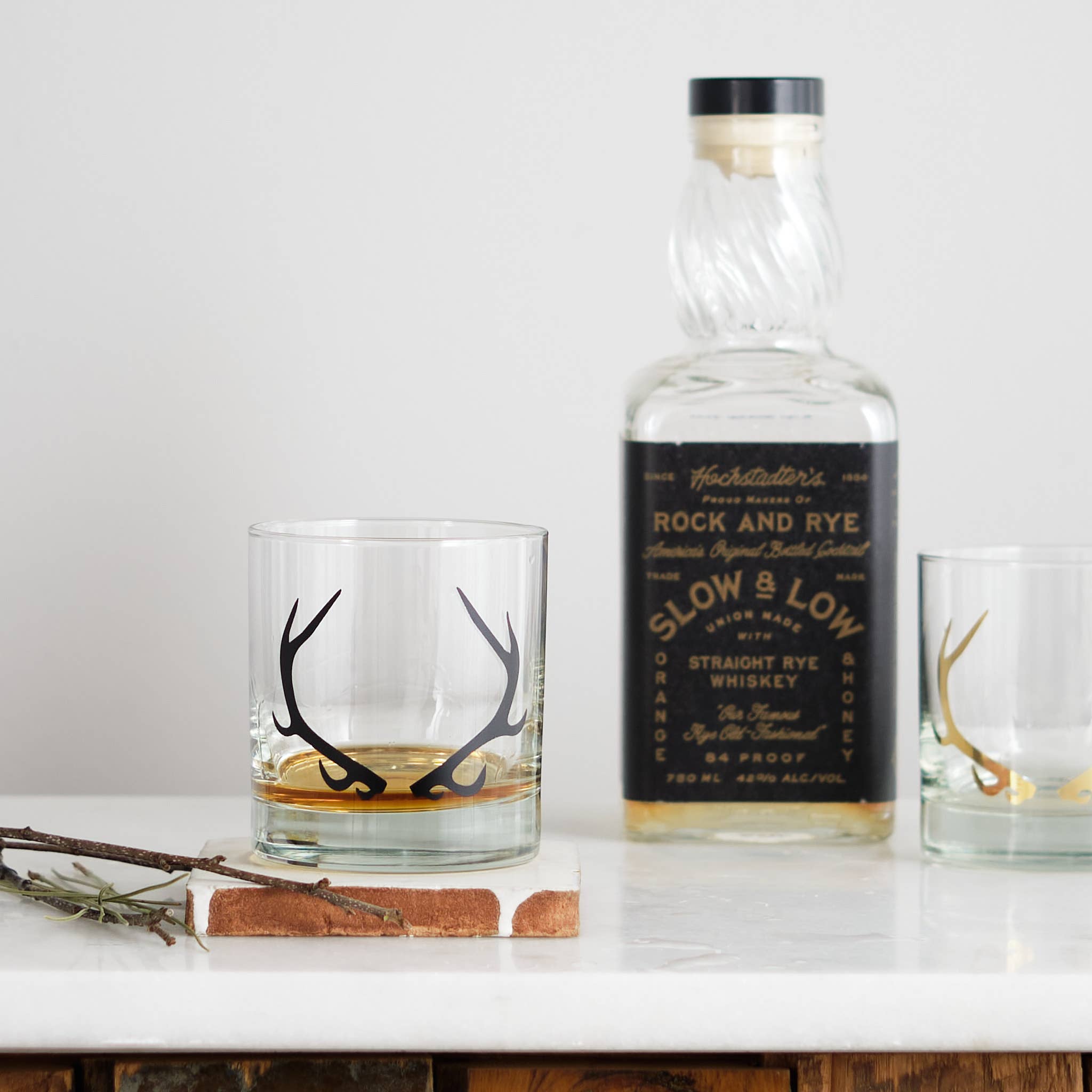 Bottle of Slow & Low Rock and Rye whiskey with two glasses on a white surface.