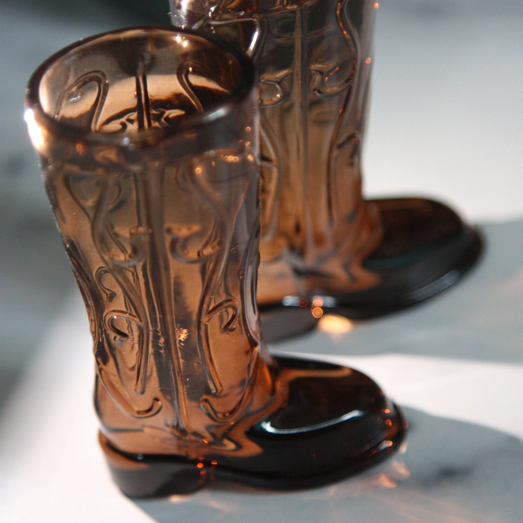 Two cowboy boot-shaped shot glasses on a reflective surface with a blurred bottle in the background.