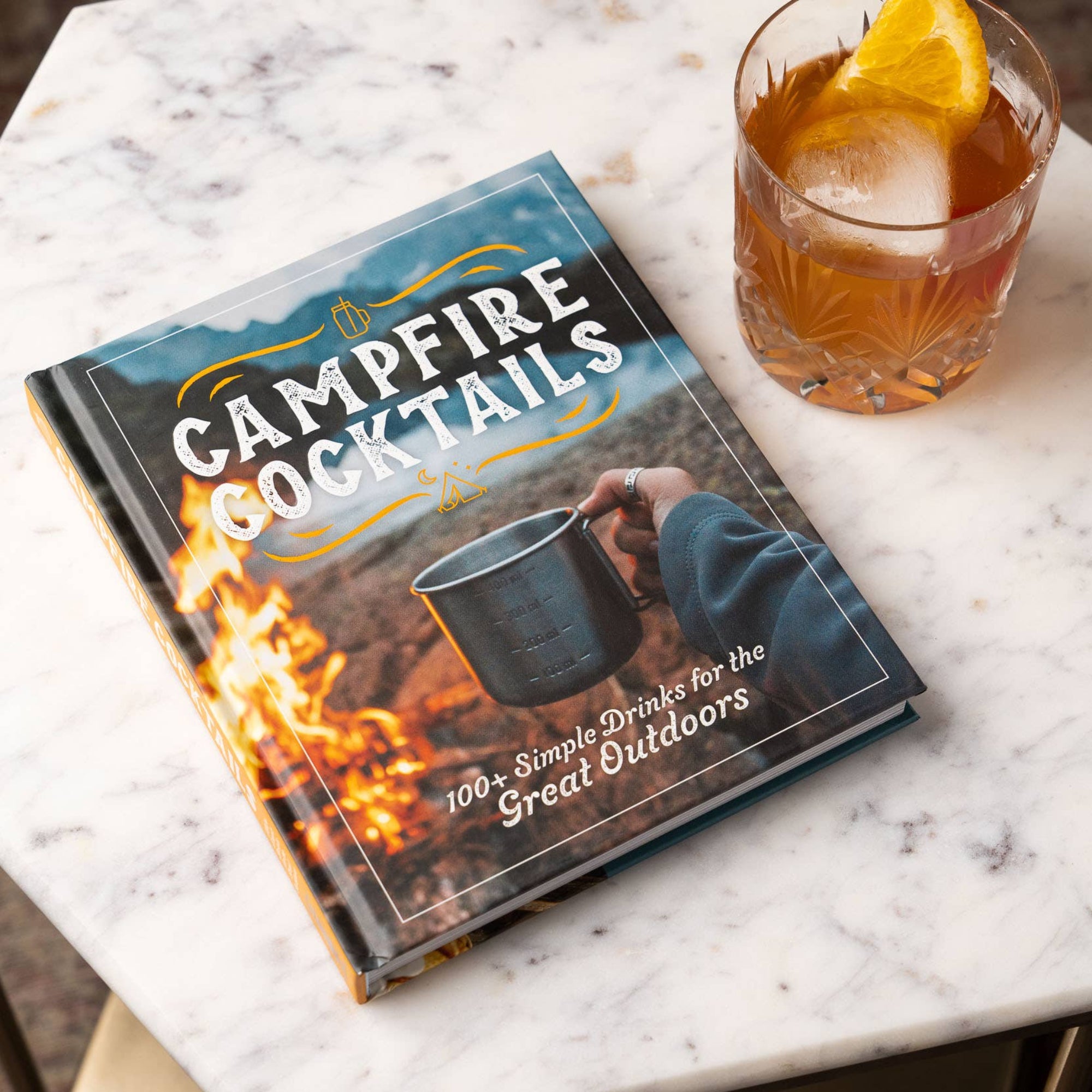 Book titled 'Campfire Cocktails' on a marble surface with a cocktail beside it.