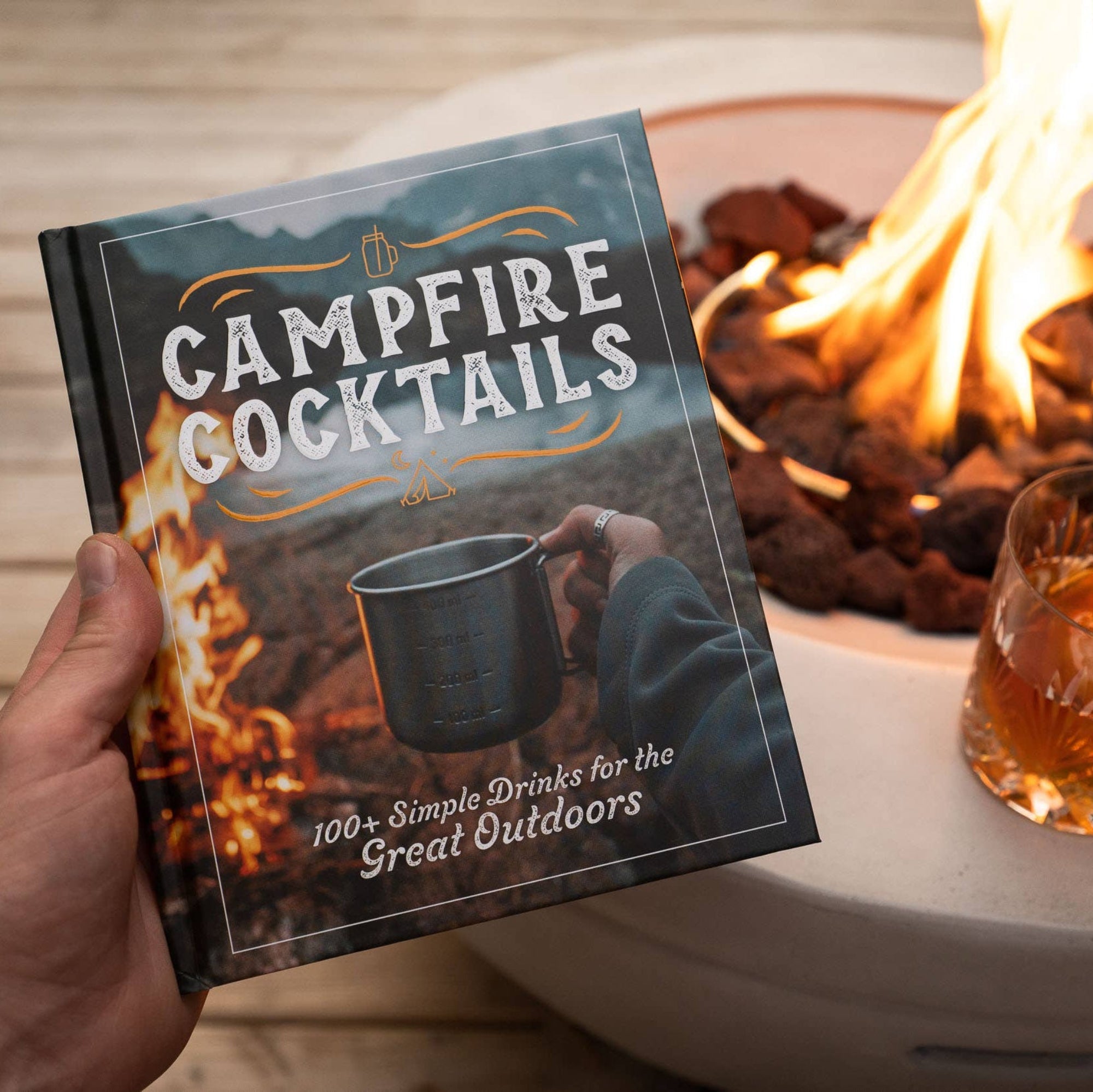 Book titled 'Campfire Cocktails' held by a hand with a cocktail in front of a campfire.