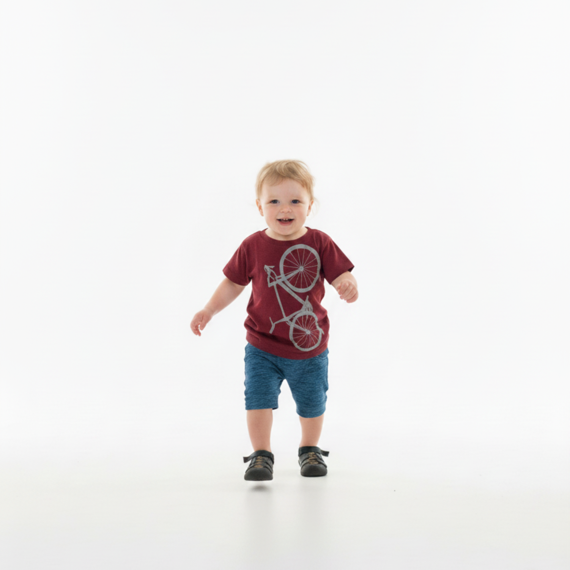 Child wearing a red t-shirt with a bicycle design, standing outdoors among greenery.