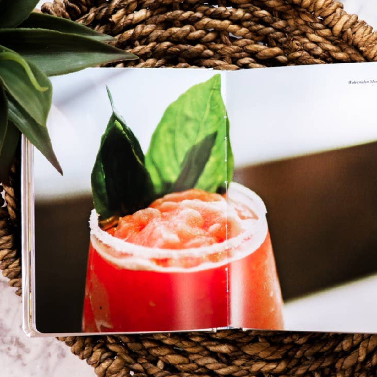 Book displaying a cocktail image on a woven coaster with a plant and candle in the background.