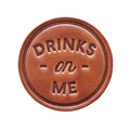 Brown leather sign with 'DRINKS on ME' text on a wooden background