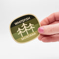 Hand holding a 'Whitefish Montana' sticker with a green and beige design.
