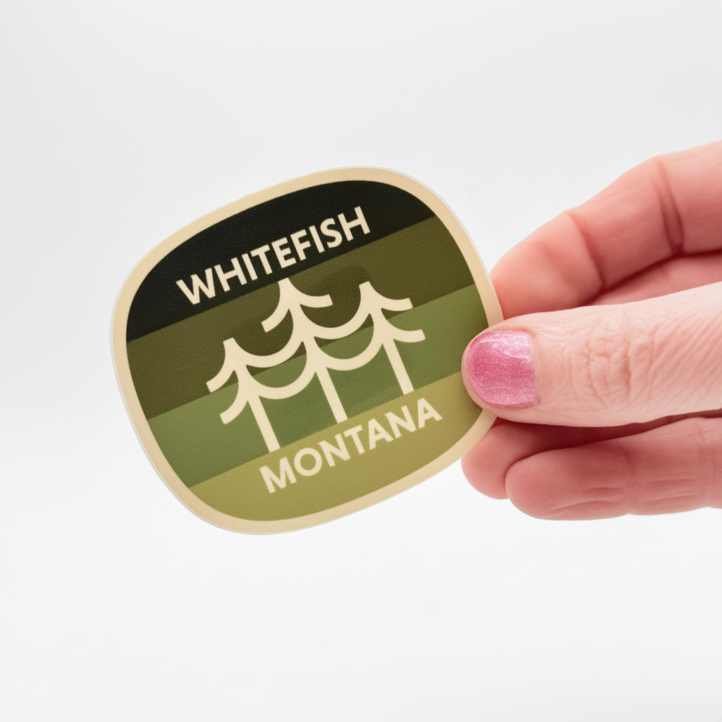 Hand holding a 'Whitefish Montana' sticker with a green and beige design.