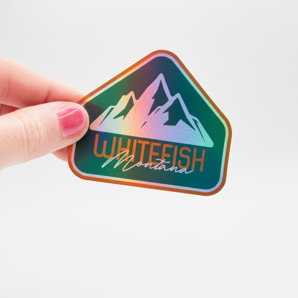 Hand holding a colorful sticker with mountain design and 'Whitefish Montana' text on a white background