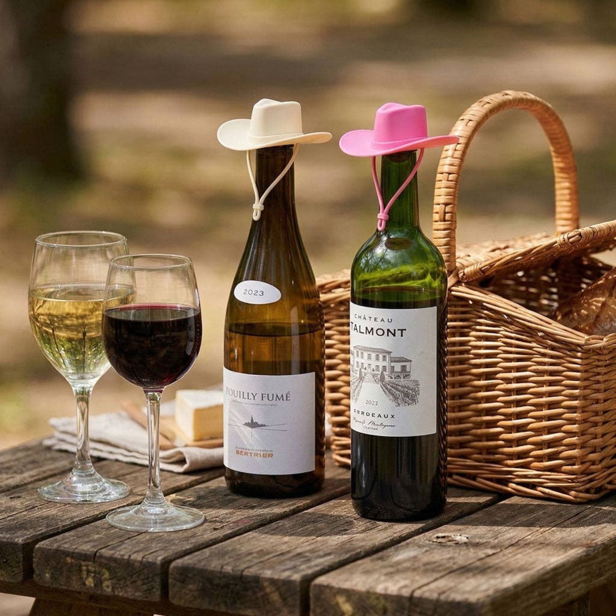 Two wine bottles with cowboy hats, two glasses of wine, and a picnic basket on a wooden table.