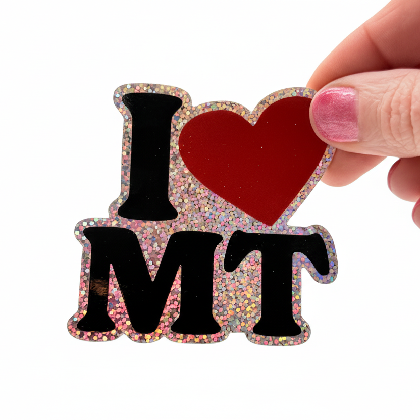 Hand holding a glittery 'I ❤️ MT' magnet on a white background