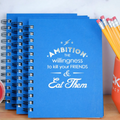 Blue notebook with sarcastic motivational quote, surrounded by pencils and a cup.