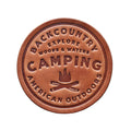 Round leather patch with 'Backcountry Camping' text and campfire graphic on a wooden background
