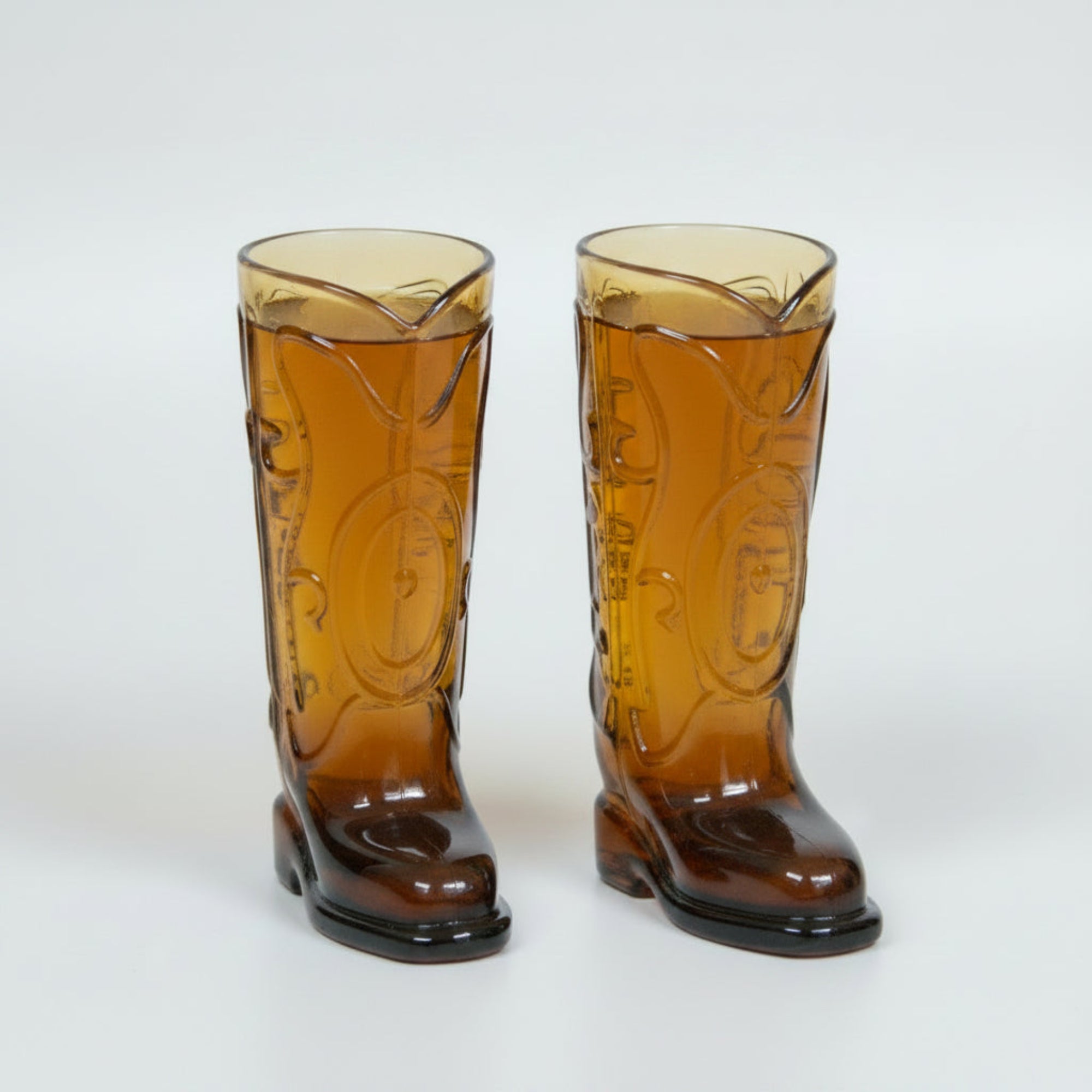 Two glass cowboy boot shot glasses on a white surface with a blurred bottle in the background.