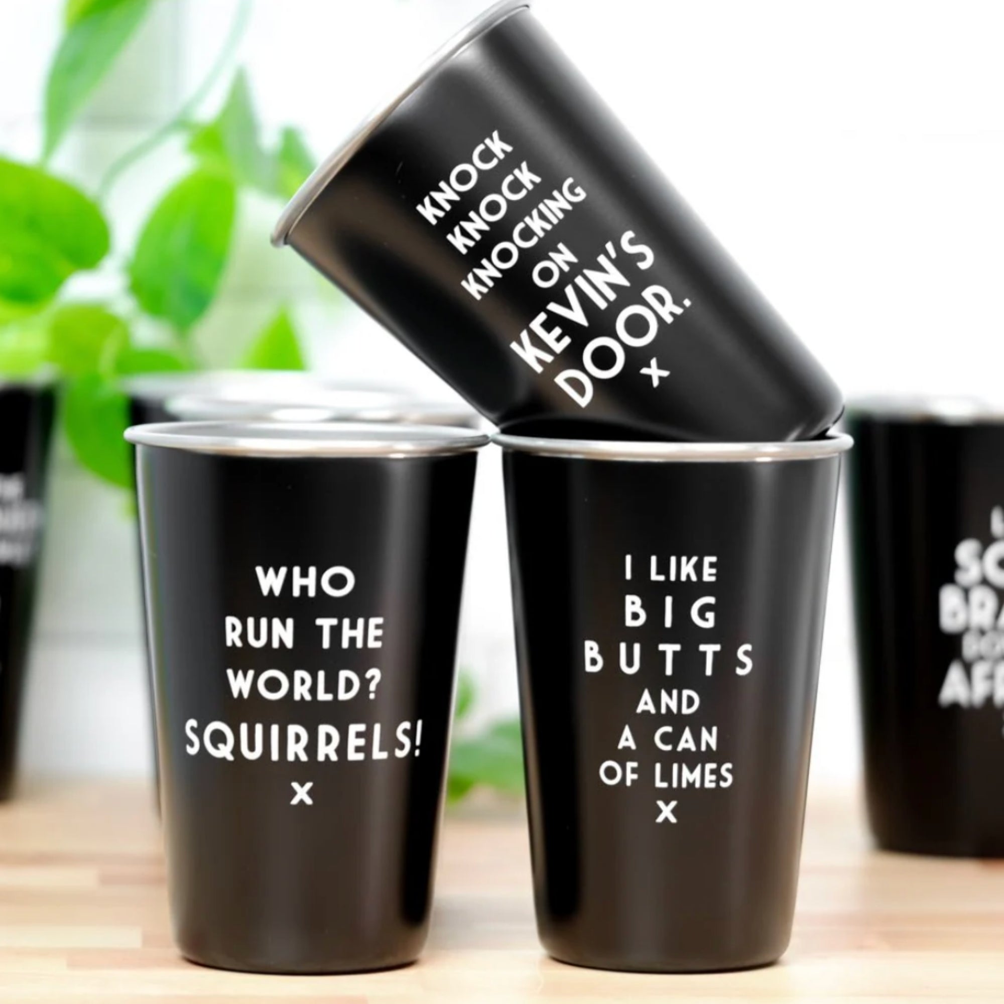 Black tumblers with humorous text on a wooden surface with plants in the background