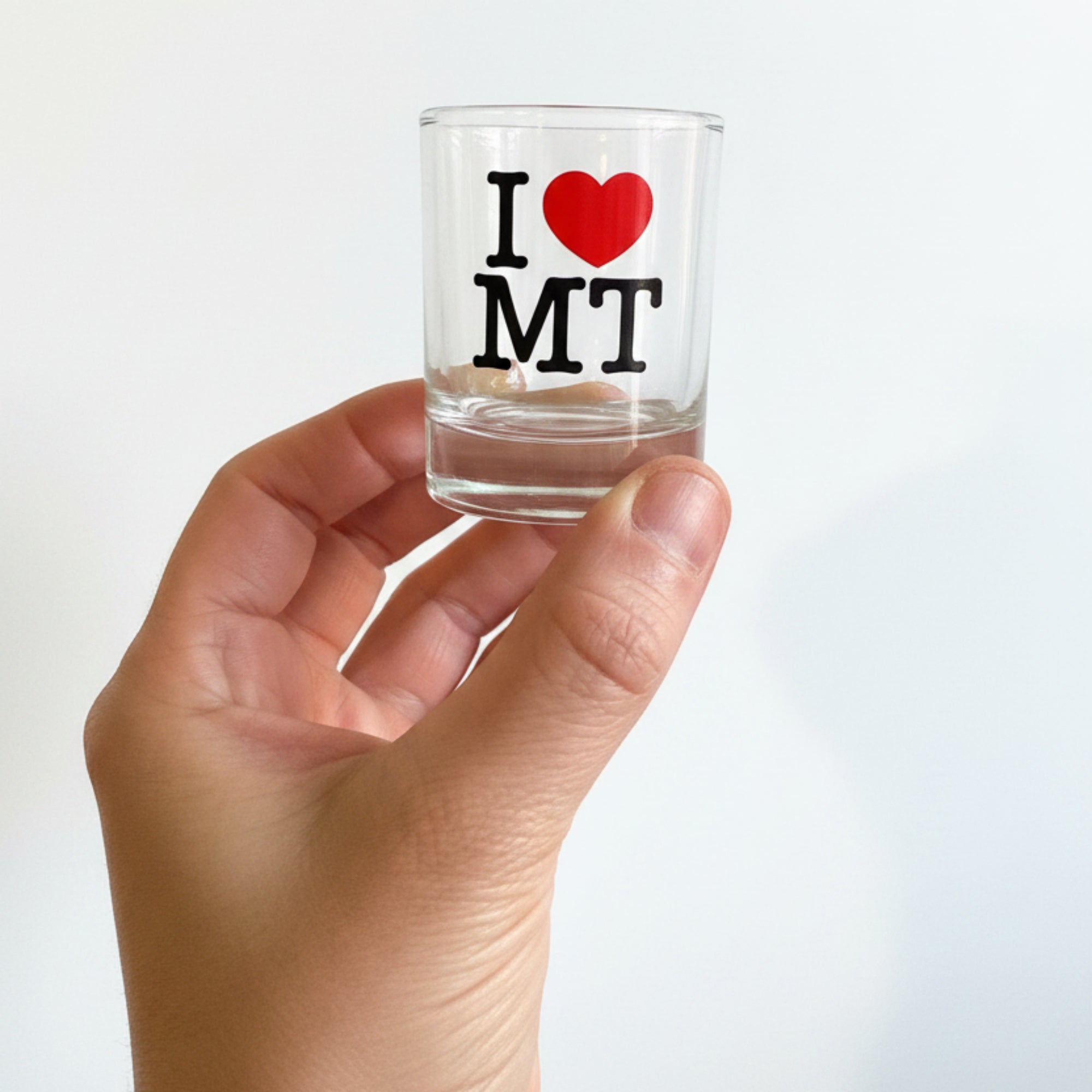 Hand holding a small clear container with 'I ❤️ MT' text against a white mesh background