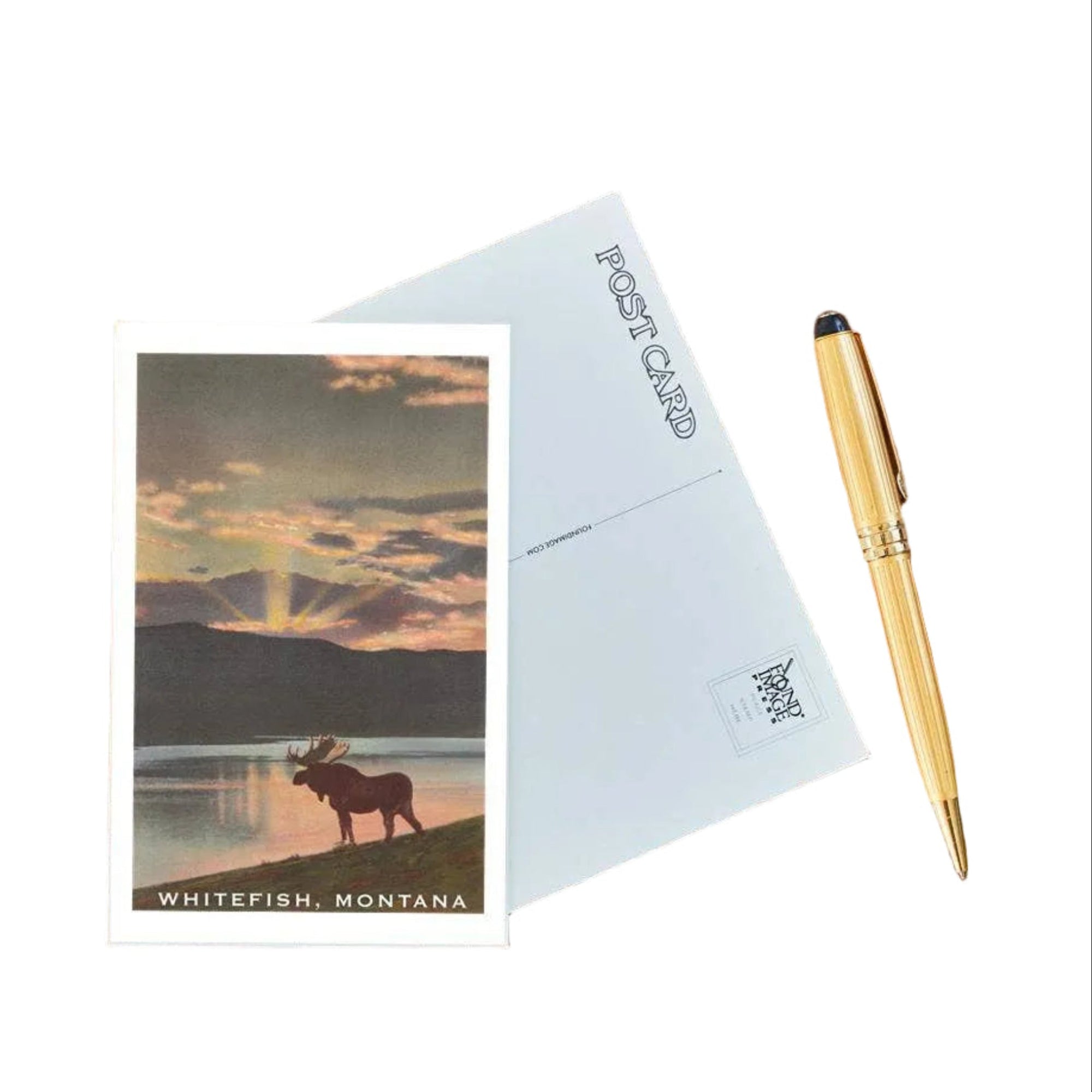 Postcard with a moose and sunset design on a wooden surface with a pen.