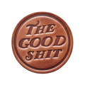 Round brown leather patch with 'The Good Shit' text on a wooden background