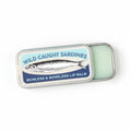 A silver metal tin of 'Wild Caught Sardines' lip balm, partially open to reveal the pale green balm inside, set against a solid light turquoise background.