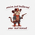 A die-cut sticker featuring a cartoon cat dressed as a cowboy with the text 'you've just buttered your last biscuit', shown on a dark wood background.