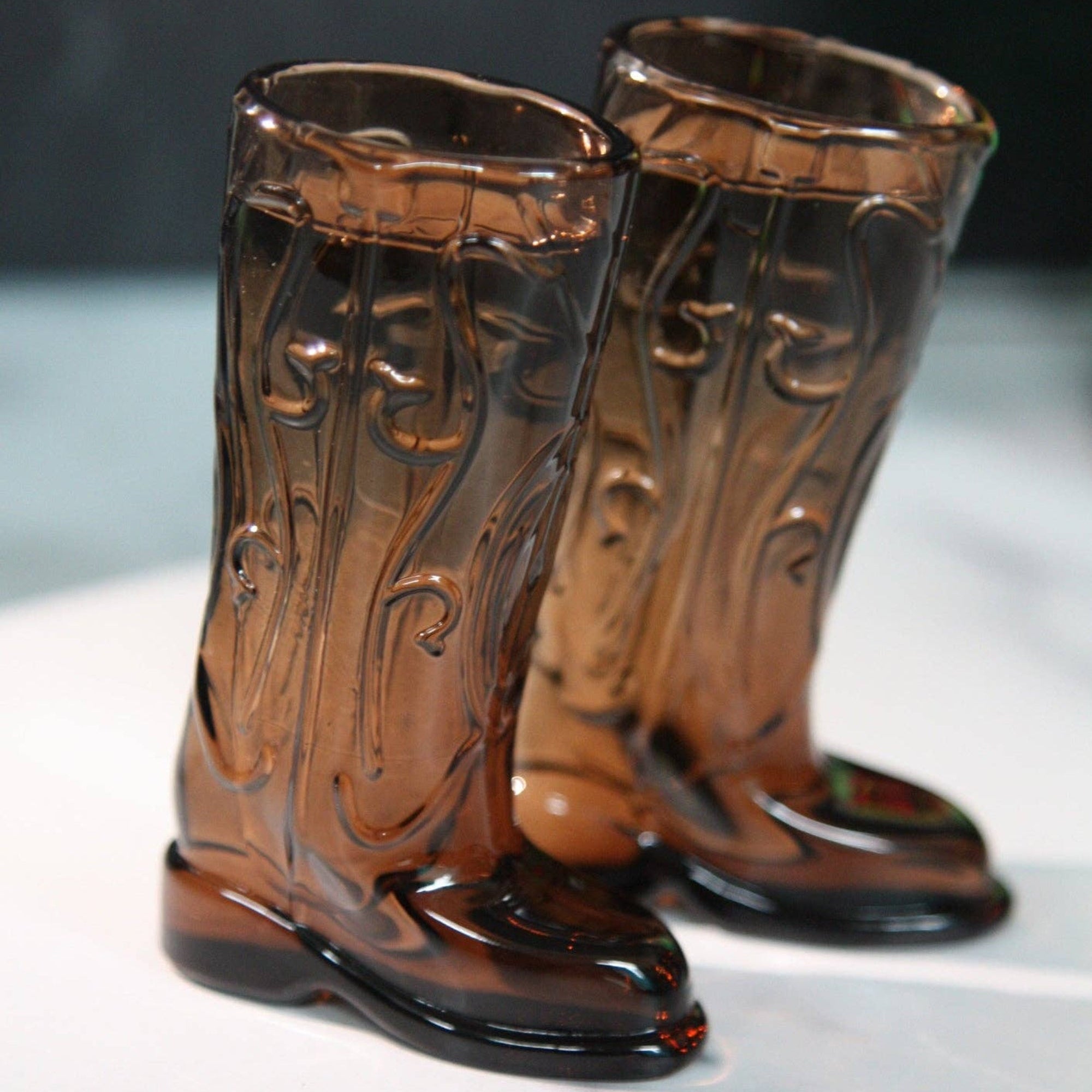 Pair of brown glass cowboy boot shapes on a white surface with a dark background
