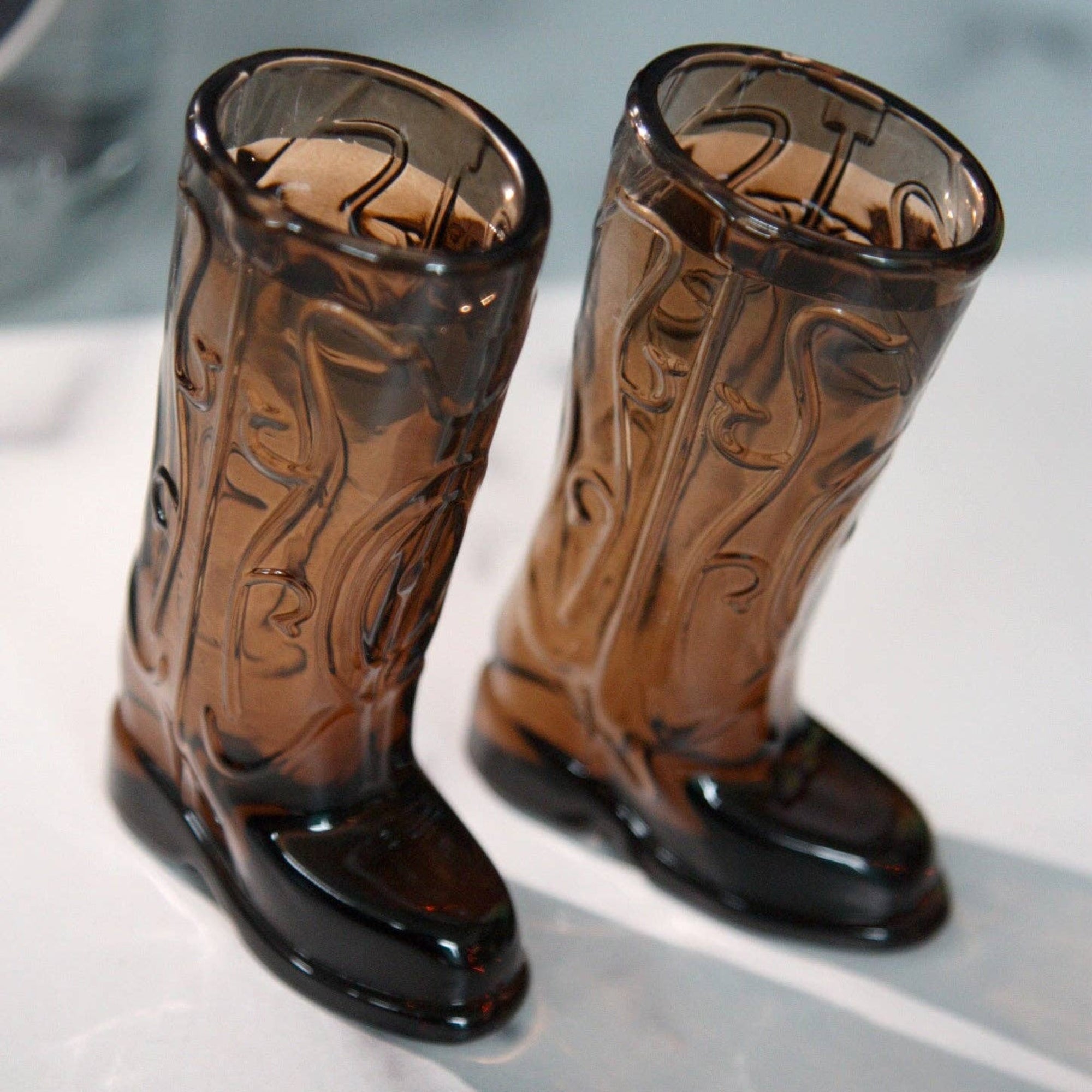 Pair of brown cowboy boot-shaped glass tumblers on a reflective surface.