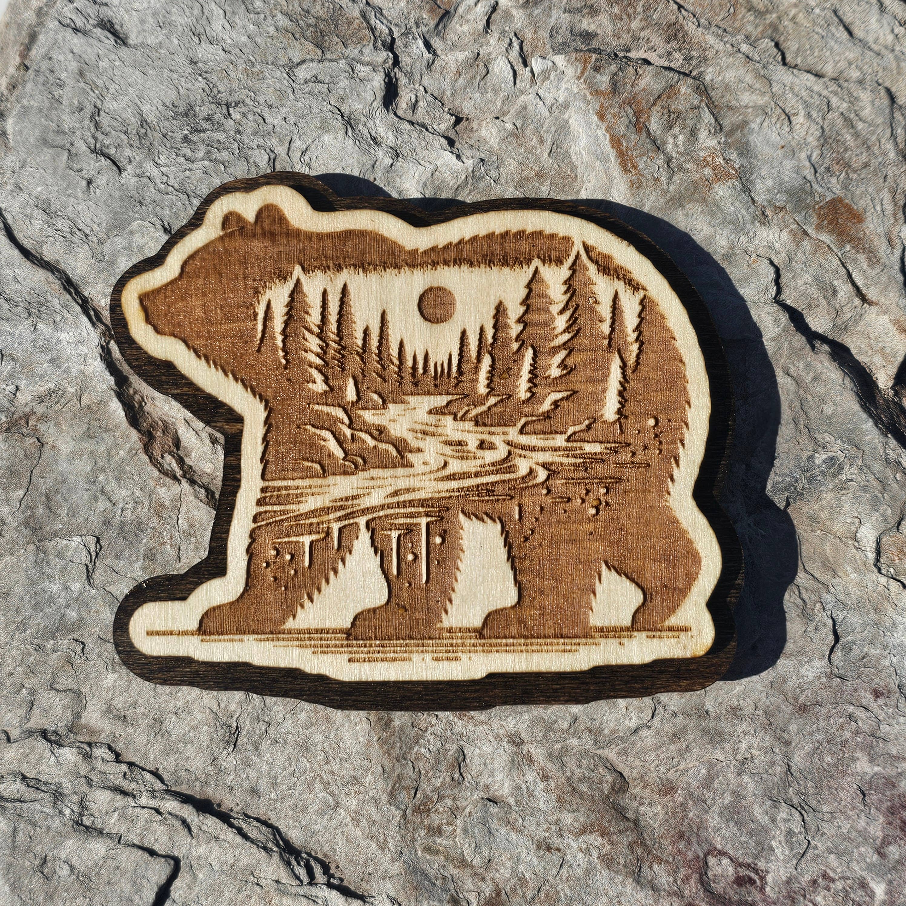 Layered wood bear magnet with an engraved forest and river scene inside the walking bear silhouette, from The Ruddy Duck in Whitefish, Montana