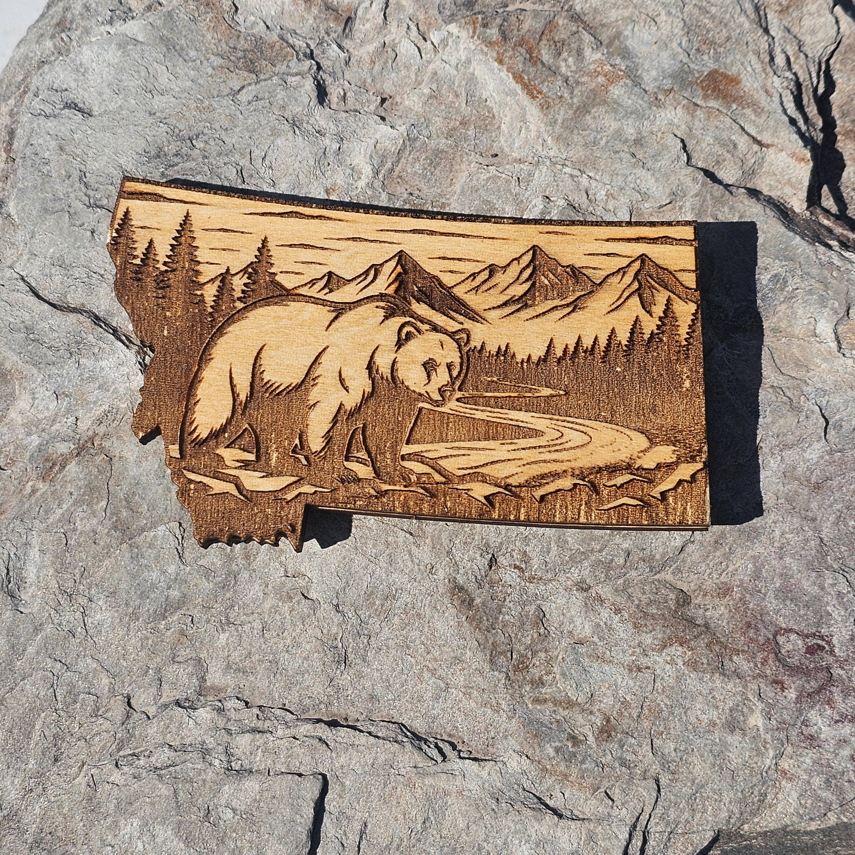 Deep engraved wood Montana state magnet showing a grizzly bear beside a winding river with mountains and pines, from The Ruddy Duck in Whitefish, Montana